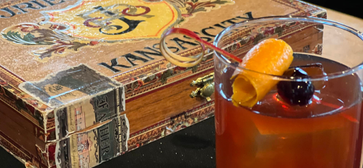 A cocktail made using Phantom V with an orange peel and cherry garnish sitting next to a Kansas City cigar box